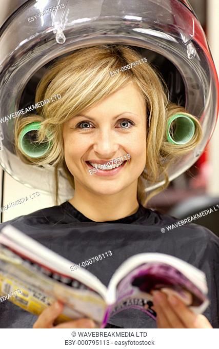 Delighted woman reading a magazine with hair curlers under a hairdryer in a hairdressing salon