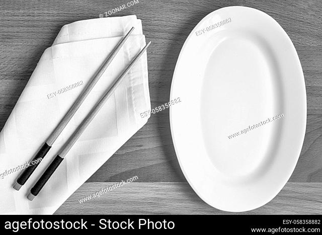 There is an empty white plate on the wooden table and wooden chopsticks on a napkin. The pattern of Japanese cuisine. The view from the top. Flat lay