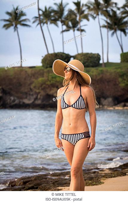 Mid adult woman wearing sunhat and bikini strolling on beach, Maui, Hawaii, USA