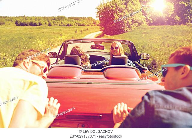 leisure, road trip, travel and people concept - happy friends pushing broken cabriolet car along country road