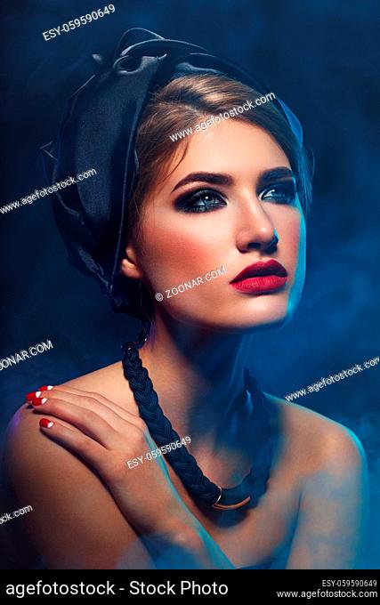 Beautiful young woman with bright makeup, red lips, black shawl on head and necklace. Studio beauty shot with blue smoke. Copy space