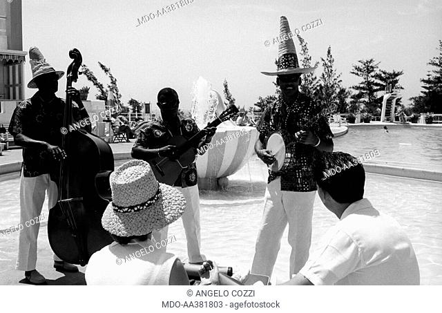 Couple on honeymoon. A married couple relaxing at the edge of a swimming pool entertained by a band of musicians. Bermuda, 1965