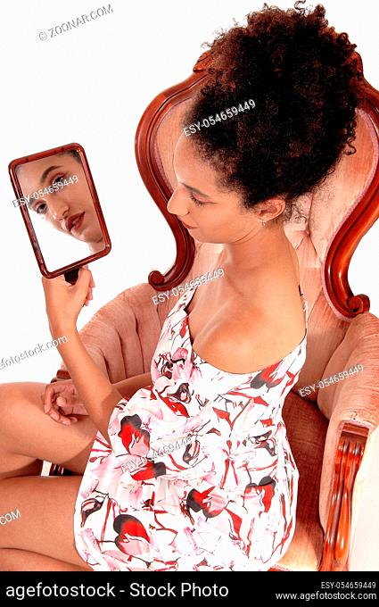 Beautiful multi-racial woman looking at herself in a hand held mirror, sitting in a summer dress and curly black hair, isolated for white background