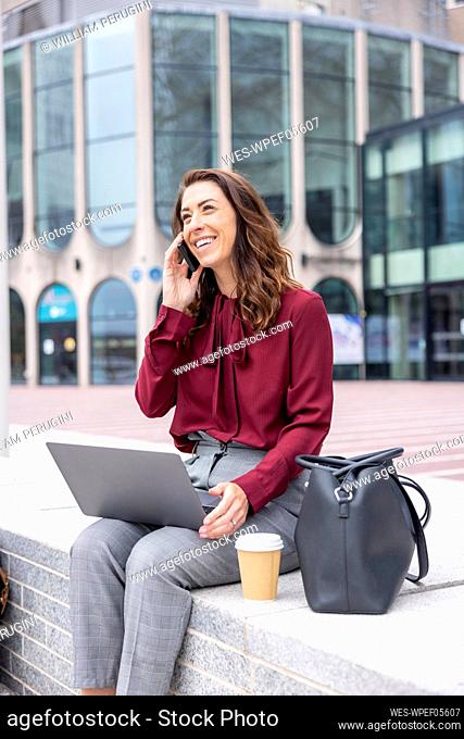 Smiling woman sitting on bench with laptop talking through smart phone at office park