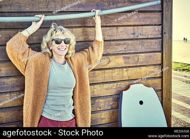 Portrait of a young mature blonde caucasian woman outdoor in a wooden house close to a beach area with a bodyboard and sunglasses