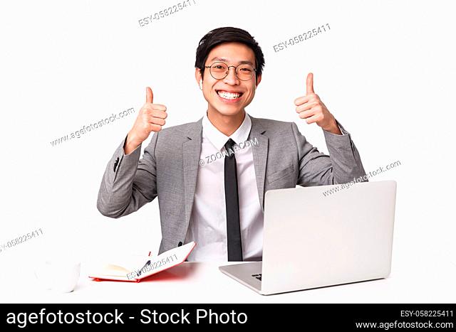 Waist-up portrait of satisfied, happy smiling asian office manager sitting at desk with laptop, notebook, drinking coffee, being productive, showing thumbs-up