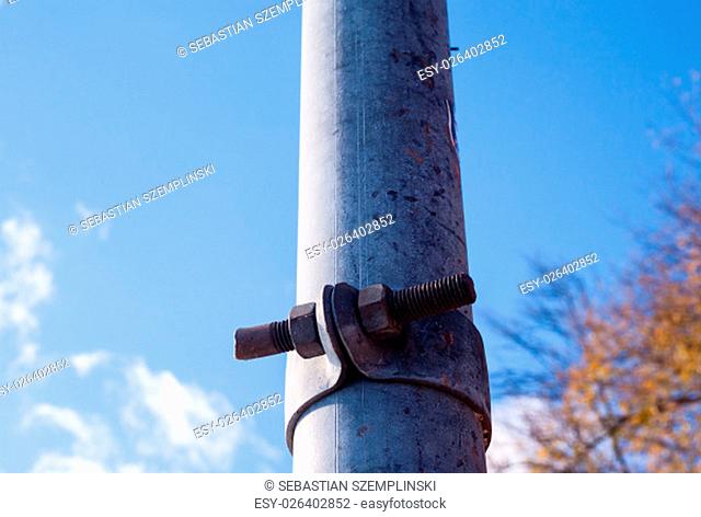 Dirty metal pole with bracket fastened with rusted screw, against blurred blue sky and branches