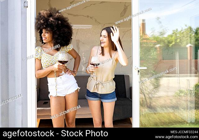 Multi-ethnic female friends waving while holding wine glass near doorway at house