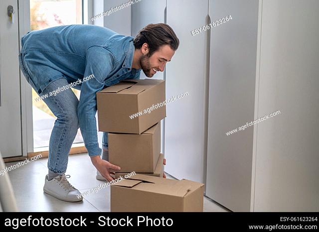 Working mood. Cheerful bearded man sideways to camera leaning over stack of boxes on floor in hallway of house