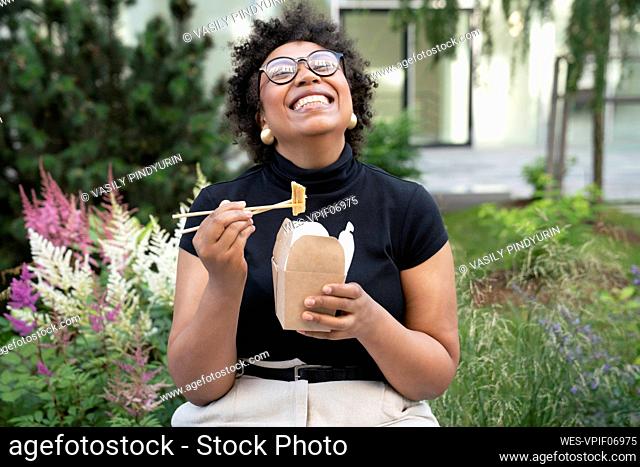 Happy young businesswoman with chopsticks and take out food sitting in office park