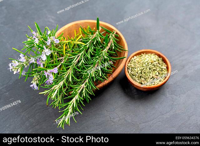 Purple blooming rosemary and fresh rosemary twigs in an olive wood bowl and a small bowl of whole dried rosemary on a black slate background