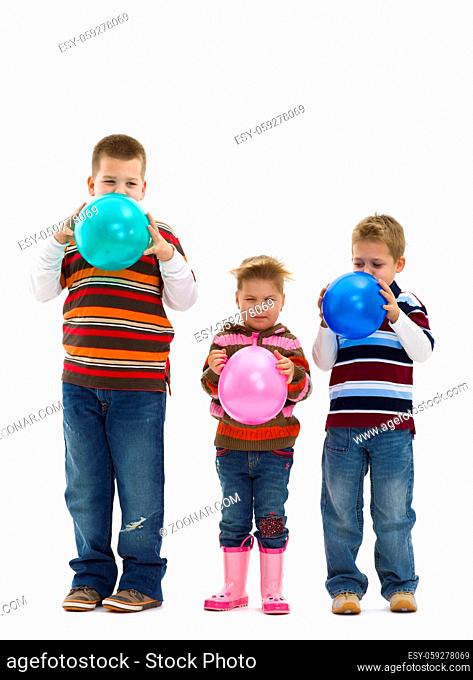 3 happy children wearing jeans and colorful striped t-shirts, blowing up toy balloons. Isolated on white background