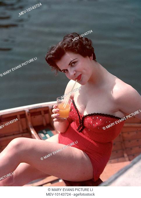 Dimple-cheeked brunette model wearing a red & black outfit of strapless, terry towelling sun-top & shorts (or bathing costume) sips orangeade as she sits in a...