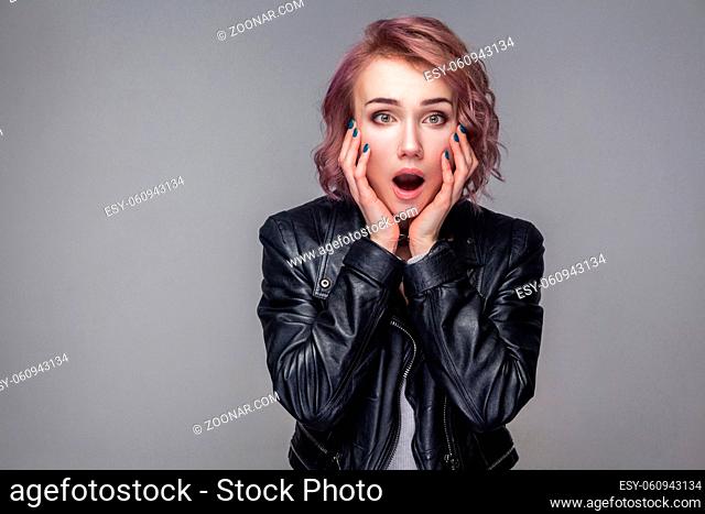 Portrait of shocked girl with short hairstyle, makeup in black leather jacket standing touching face and looking at camera with big eyes and open mouth