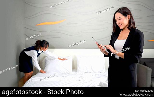 Asian businesswoman in black suit standing with tablet computer in her hand. The hotel manager verify the tidiness of the room for hotel guests while the maid...