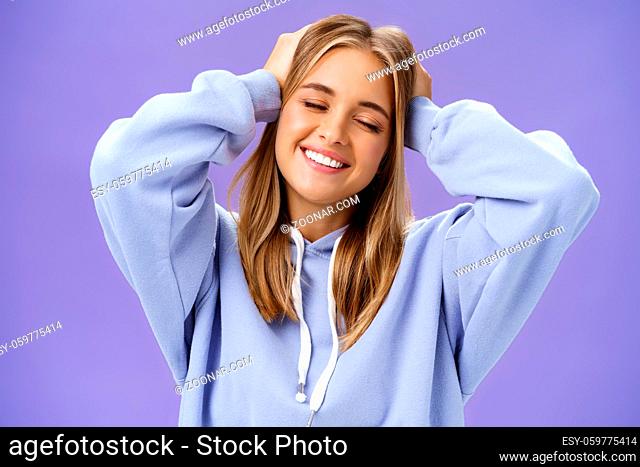 Tender relaxed charming woman with blond hair touching head joyfully smiling broadly with closed eyes daydreaming posing in trendy hoodie against purple wall...