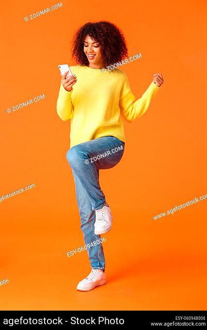 Full-length vertical shot cheerful cute technology addicated african-american modern girl with afro haircut, jumping and triumphing as reading great news from...