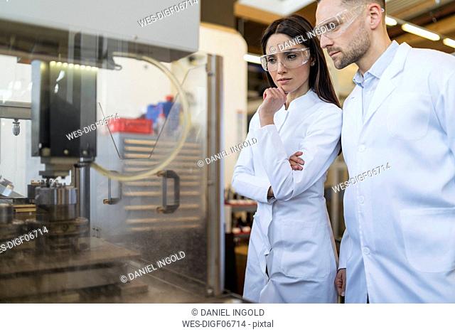 Colleagues wearing lab coats and safety goggles looking at machine in modern factory