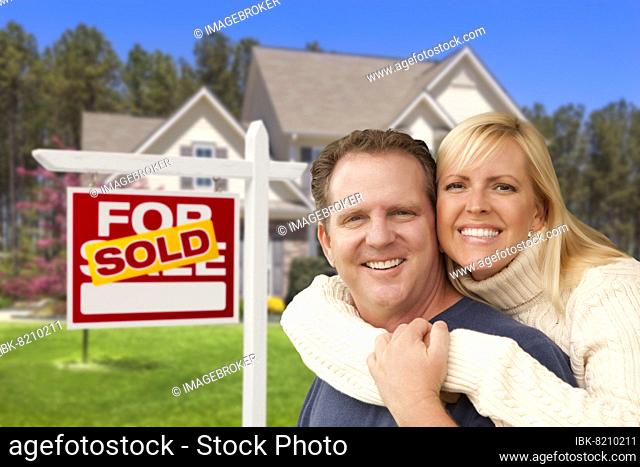 Happy couple hugging in front of sold real estate sign and house