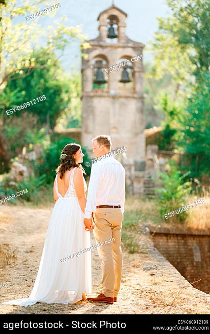 Bride and groom walk to the ancient bell tower near the church in Prcanj holding hands . High quality photo