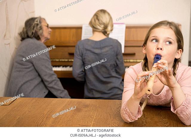 Girl playing recorder while sister on piano watched by grandmother