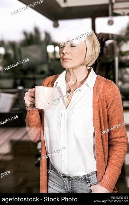 Blond businesswoman with eyes closed holding coffee cup seen through glass