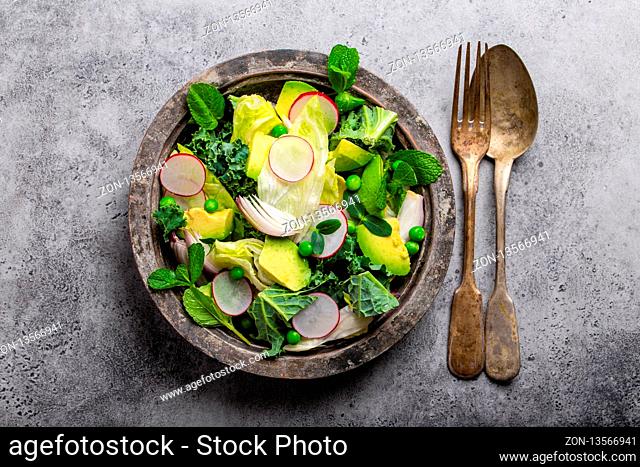 Green organic vegetables salad with avocado, kale, green peas, sprouting herbs, radish in rustic bowl on gray concrete background, close-up, top view