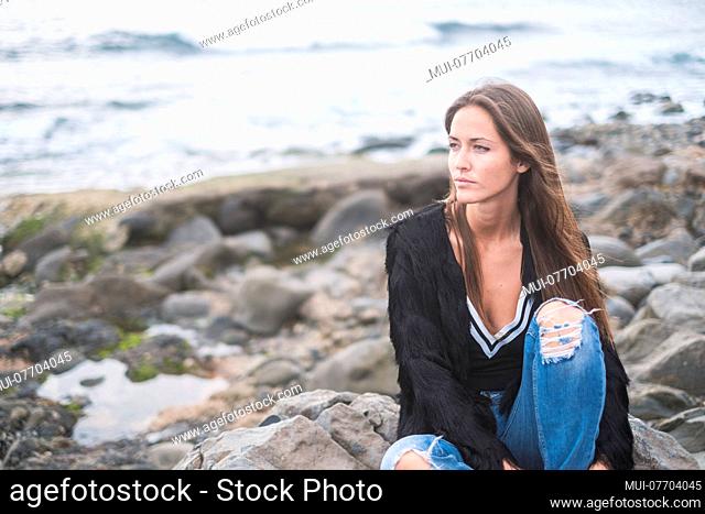 Indie young caucasian woman enjoy the nature and the freedom alone on the rocks at the beach, ocean in background, casual clothes and wanderlust concept