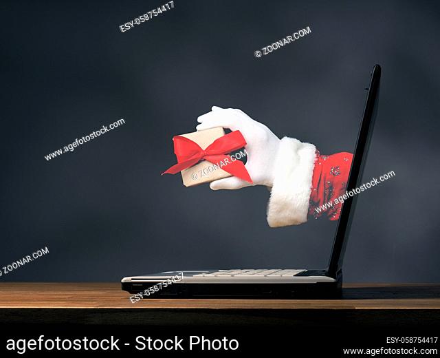 Hand of Santa is holding present with a red bow. Open laptop on a wooden table, Christmas shopping or internet shopping concept