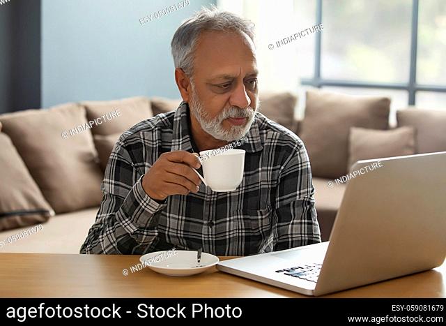 A SENIOR ADULT MAN DRINKING TEA WHILE LOOKING AT LAPTOP