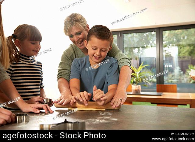 Happy woman helping son in rolling dough by daughters at home