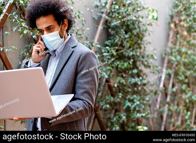 Afro businessman wearing protective face mask holding laptop while talking on smart