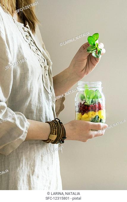 A woman in a linen dress holding a jar of fruit salad with mango, pomegranate seeds and lambs lettuce