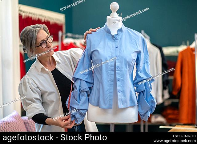 Reflections. Middle-aged woman in glasses with measure touches sleeve of blouse thoughtfully looking at dummy standing in atelier