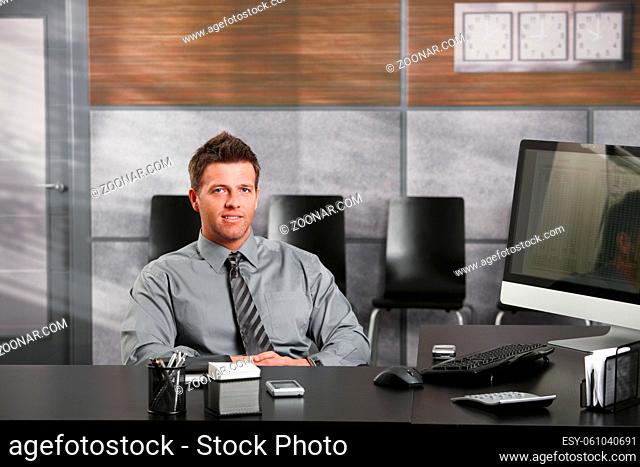 Portrait of happy businessman sitting at office desk, looking at camera, smiling