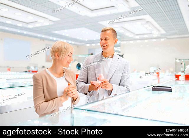 Love couple buying gold decoration. Male and female consumers looking on jewels in jewelry store. Man and woman choosing wedding rings