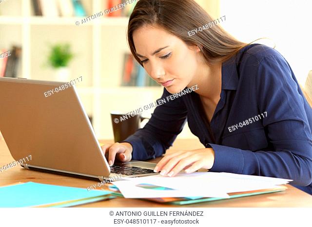 Concentrated entrepreneur comparing documents on line with a laptop sitting in a desktop at home