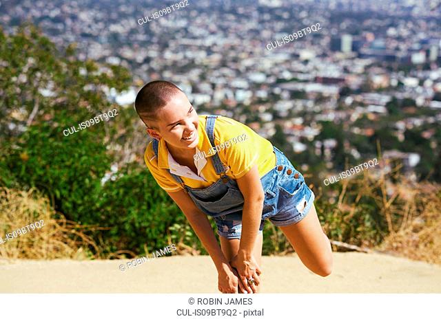 Teenage girl with cropped hair balancing on one leg on cityscape hilltop, Los Angeles, California, USA