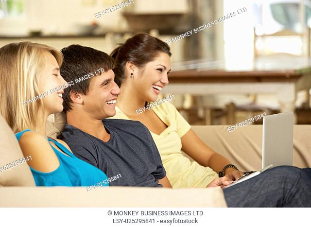 Three Teenagers Sitting On Sofa At Home Using Tablet Computer And Laptop Whilst Watching TV