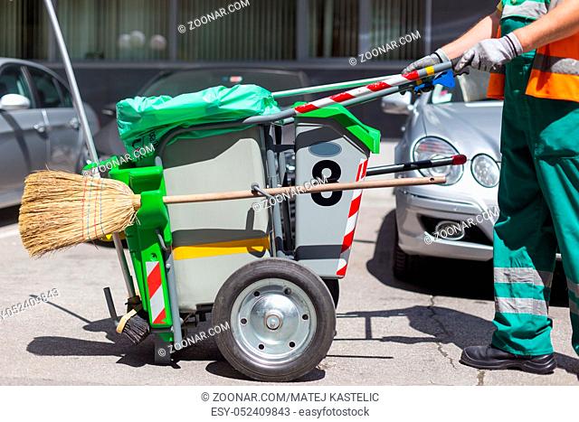 Worker of cleaning company in green uniform with garbage bin and a broom. Utility service company men worker of municipality sweeping city streets