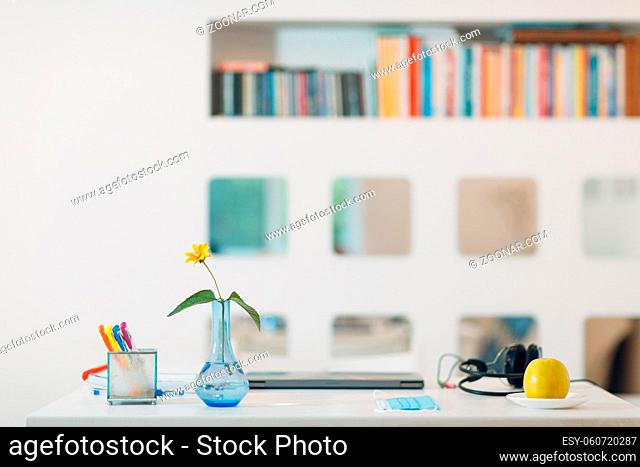 Modern house interior workspace with a silver laptop, headphones, vase, flower, apple, face mask and pens on the table with white wall background and book shelf
