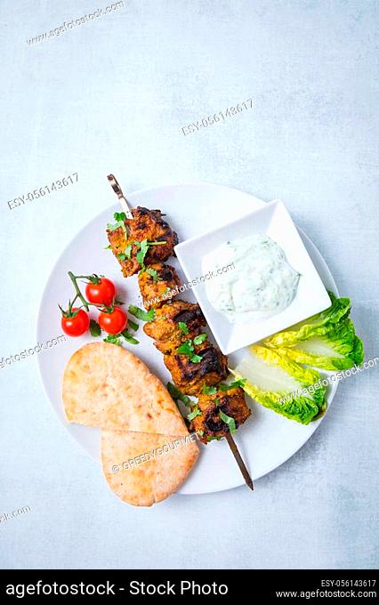 Top view of lamb tikka bbq skewer with fresh parsley, pita bread, tomatoes, lettuce and sauce on a white plate. Light background