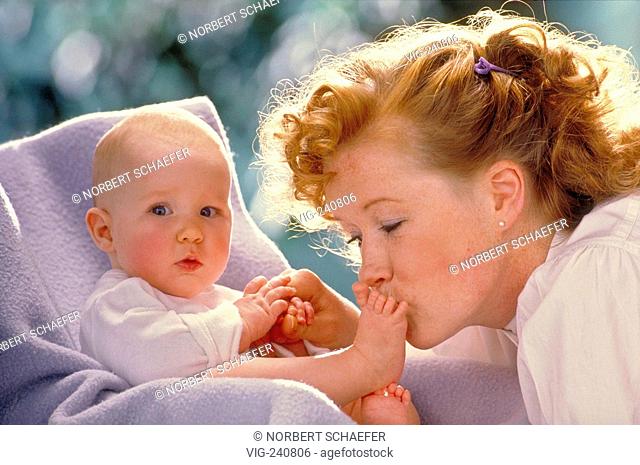 portrait, close-up, freckeled woman with red curly hair kisses tenderly the naked feet of her baby sitting on a lilac blanket in a childrens seat  - GERMANY