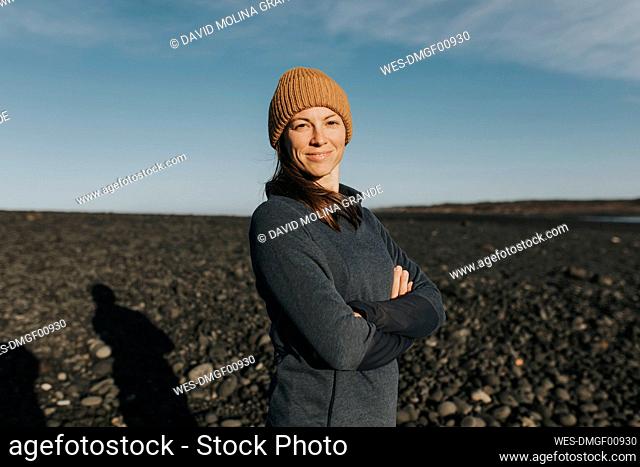 Happy woman wearing knit hat standing with arms crossed at Janubio Beach, Lanzarote, Canary Islands, Spain