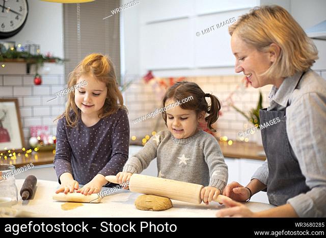 Blond woman wearing blue apron and two girls standing in kitchen, baking Christmas cookies