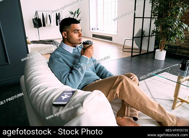 Man holding disposable coffee cup sitting by smart phone with flight mode symbol on sofa at home