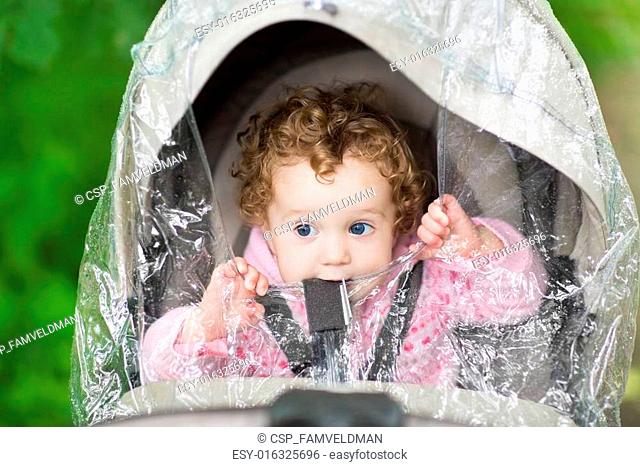 Cute curly baby girl sitting in a stroller under a plastic rain