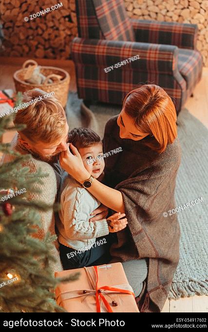Theme family holiday New Year and Christmas. Young caucasian family mom dad son on wooden floor near fireplace christmas tree on Christmas evening
