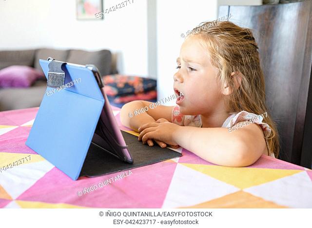 portrait of four years old blonde cute girl watching digital tablet on table and complaining face expression, sitting in living room of home