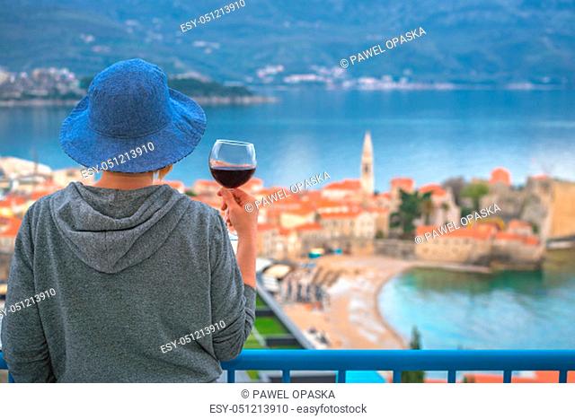 Caucasian woman wearing blue hat standing in an apartment balcony and holding glass with red wine looking at the Budva town below, Montenegro